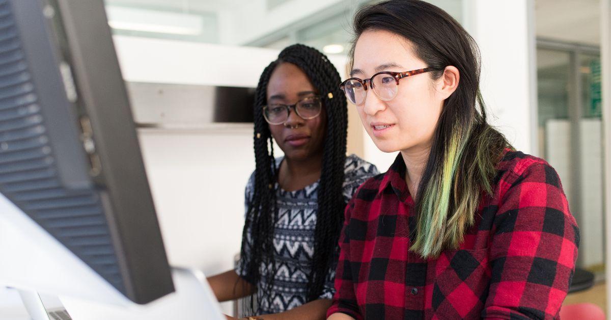 two women sit beside each other at a desk, looking at the same monitor, pair programming